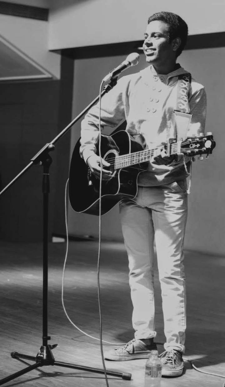 Rameshwar smiling on stage with an acoustic guitar and microphone stand, in black and white