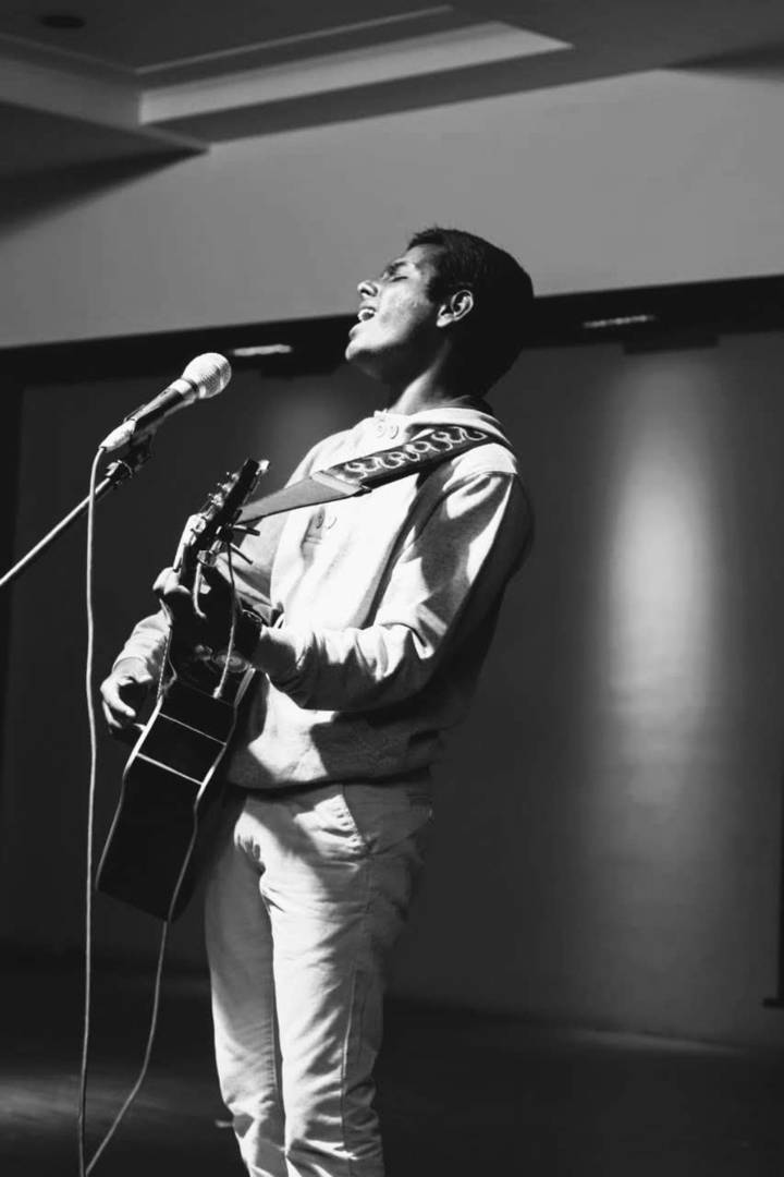 Rameshwar singing into a microphone while playing an acoustic guitar, eyes closed, in black and white